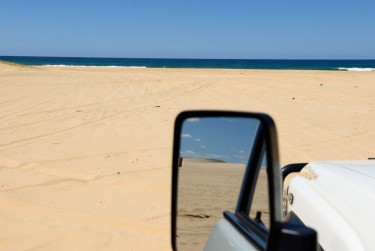 View of the beach from the car, with the sand dunes and blue sky in the side mirror